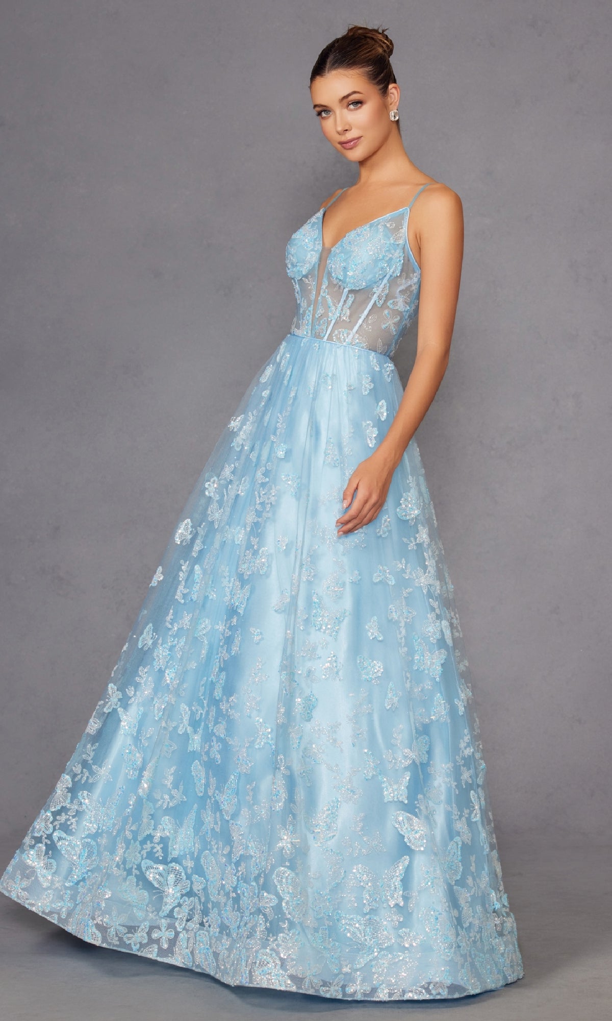 A woman models the Sheer-Corset Glitter-Print Long Prom Dress 2413, featuring a light blue A-line silhouette with thin straps, a fitted bodice, sparkling details, and a sheer overlay against a plain gray backdrop.